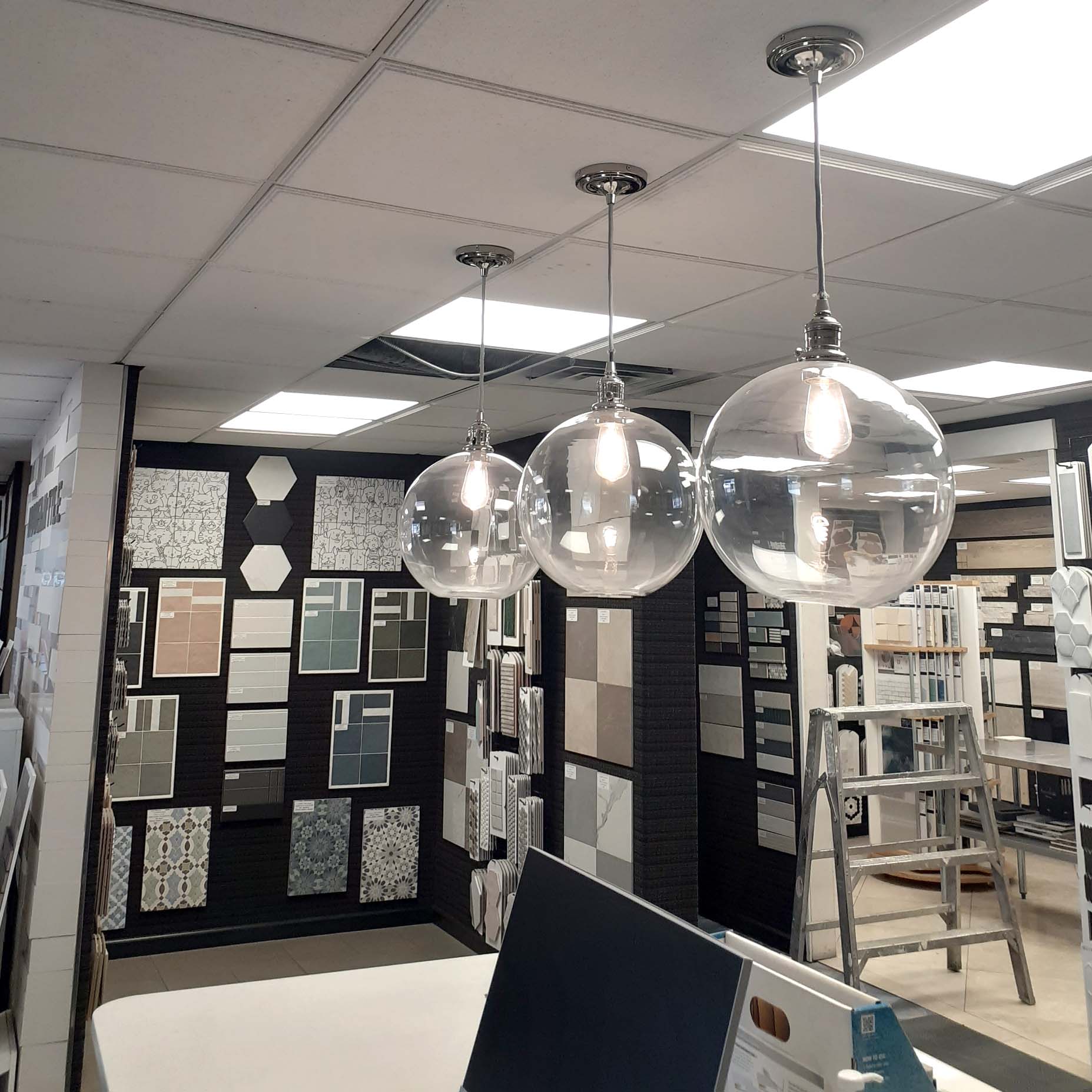 Three clear globe pendant lights hang from the ceiling in a showroom displaying various tile samples on the walls, with a metal ladder in the background.