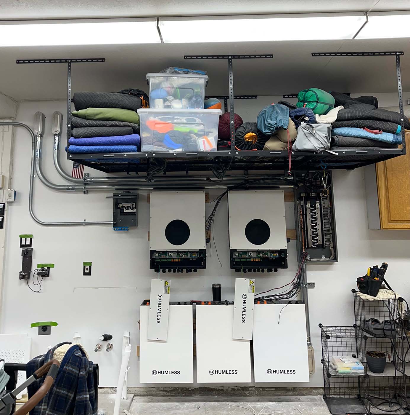 A garage wall with energy storage units, electrical panels, and a ceiling rack holding bins, camping gear, and blankets—perfectly organized for generator installation or backup power solutions.
