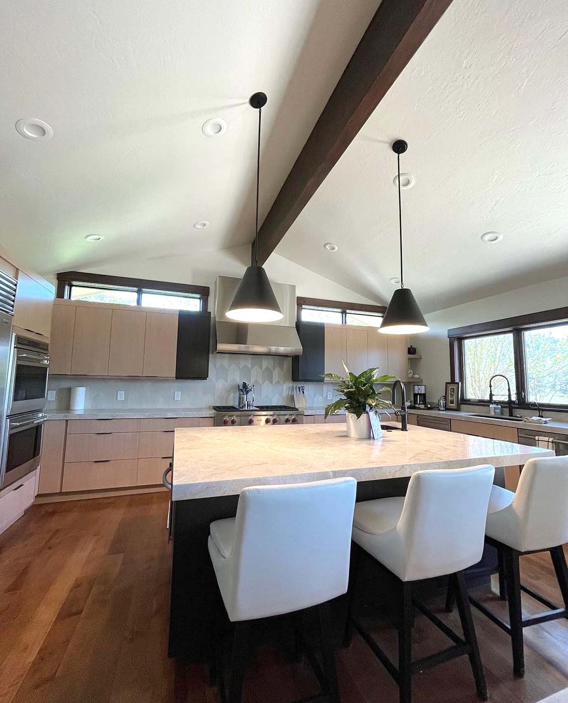 Modern kitchen with light wood cabinets, black accents, a large island with white chairs, pendant lights, and stainless steel appliances—expertly designed with recent electrical remodeling to enhance both functionality and style.