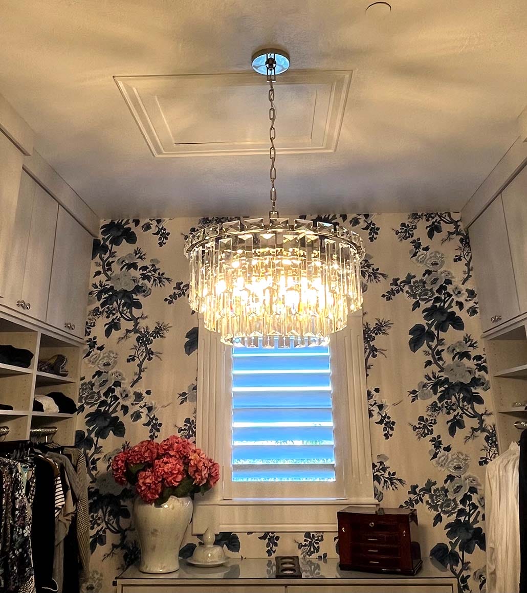 A stunning crystal chandelier, expertly showcased by a flawless lighting installation, hangs from the ceiling in a walk-in closet adorned with floral wallpaper. Shelves lined with clothing, a window draped with shades, and a vase of pink flowers on the counter complete this elegant retreat.