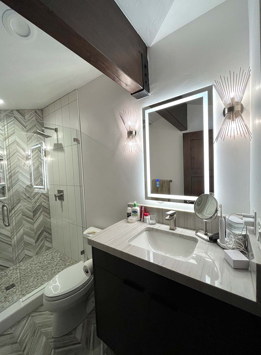 Modern bathroom with a glass shower, backlit mirror, wall sconce lights, dark vanity, and toiletries on the marble countertop—featuring thoughtful electrical remodeling to seamlessly integrate contemporary lighting and smart fixtures.