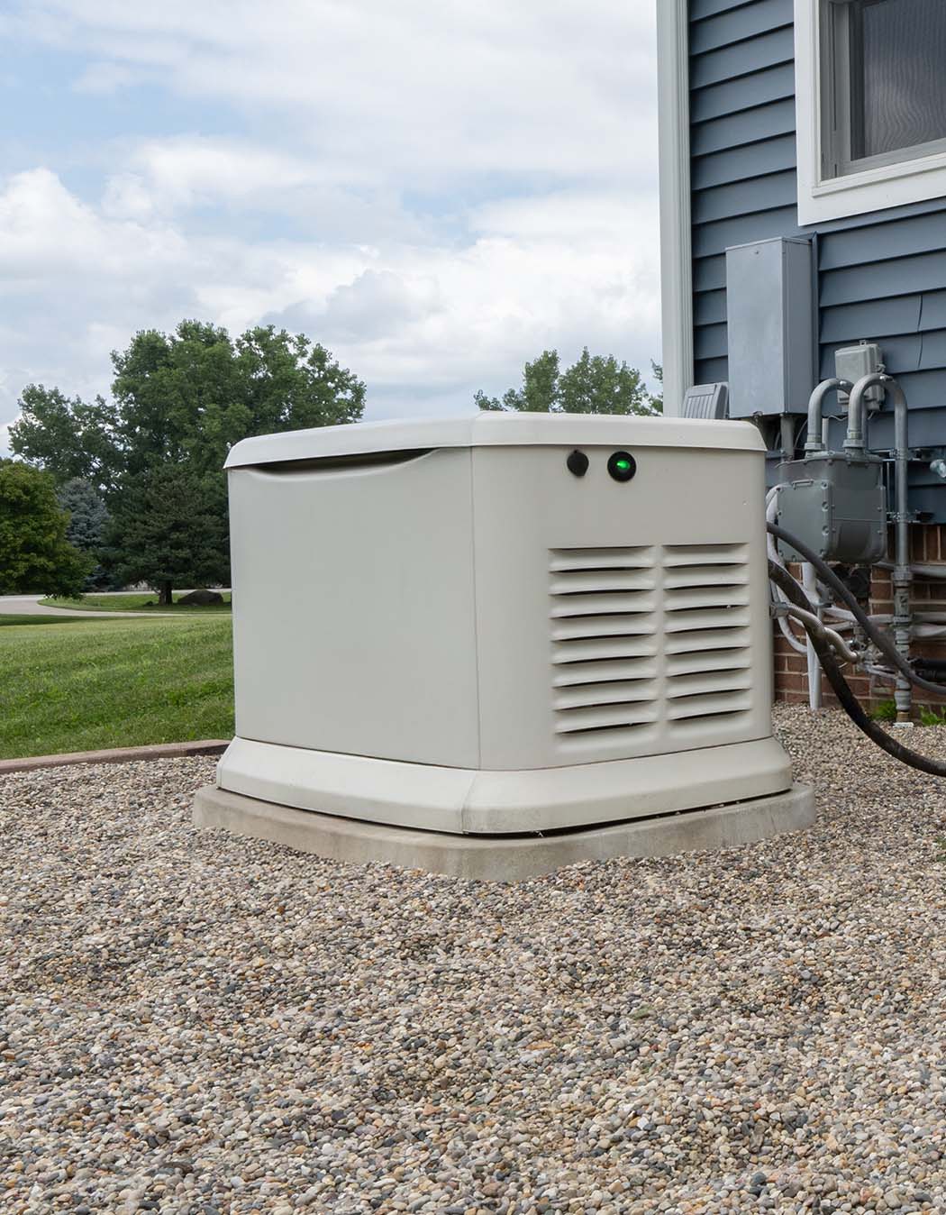 A backup generator is installed on a gravel pad outside a house with siding, connected to utility lines.