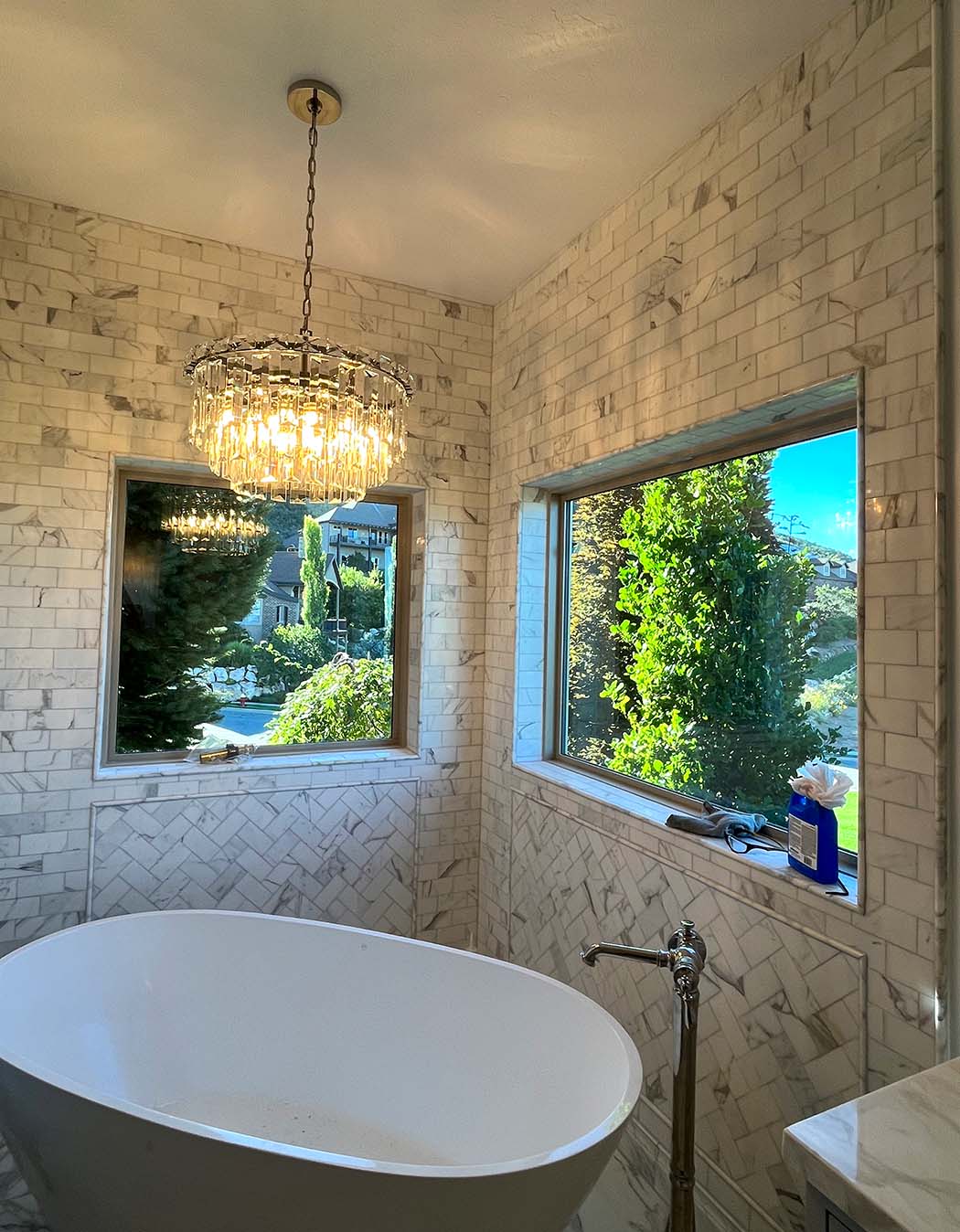 A bathroom with a freestanding tub, marble-patterned tile walls, two large windows, a modern chandelier, and a tub faucet.